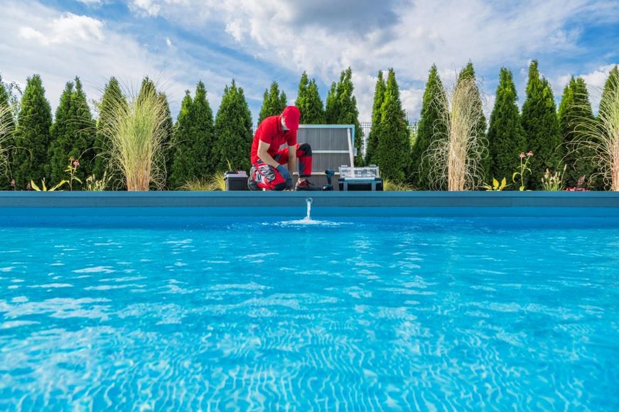 Person in red shirt working on pool equipment next to a swimming pool, with trees and sky in the background.