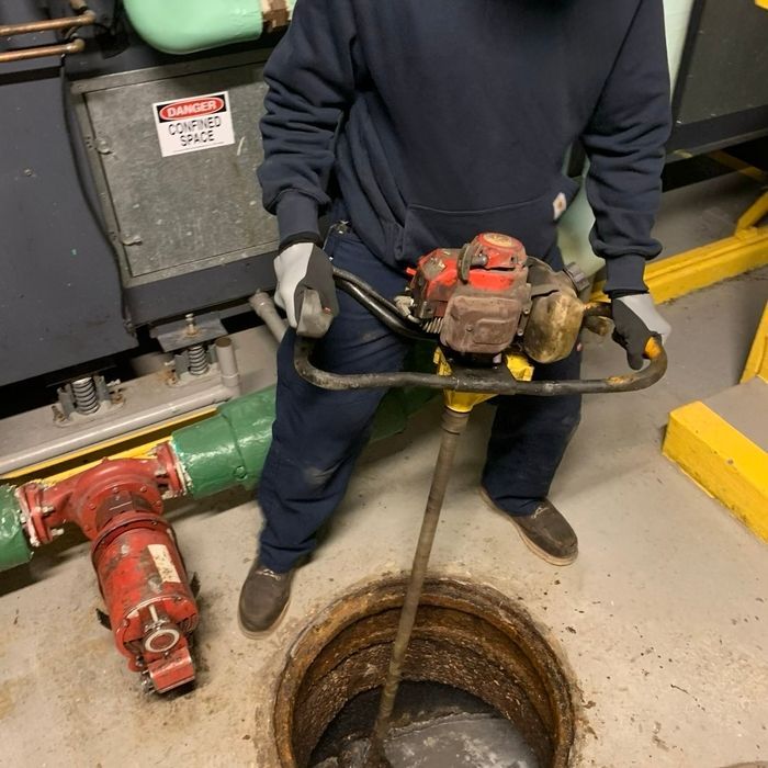 Worker in dark coveralls lowering a pump into an open manhole in an industrial area.