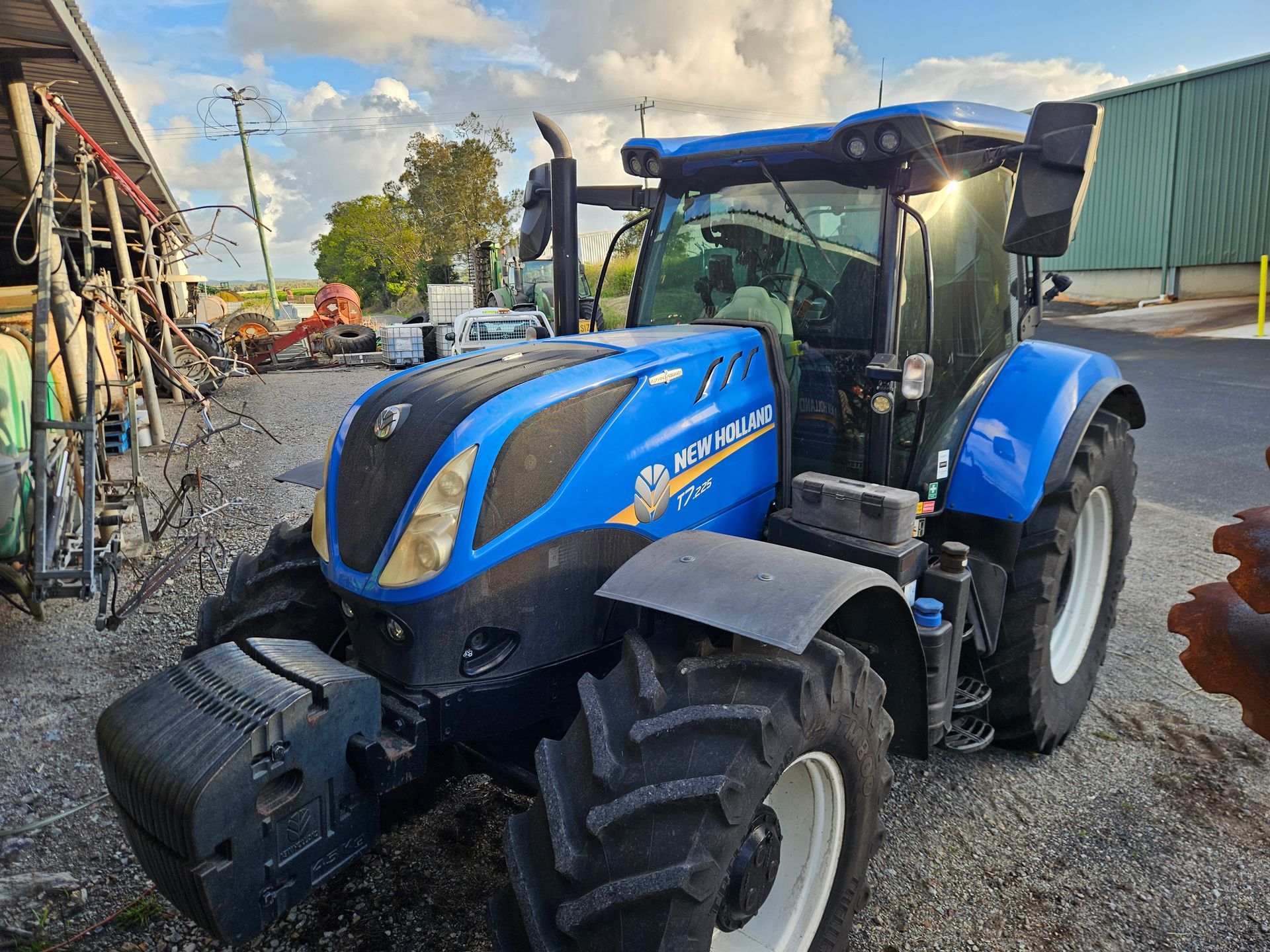 Blue New Holland tractor parked on gravel beside a barn on a sunny day