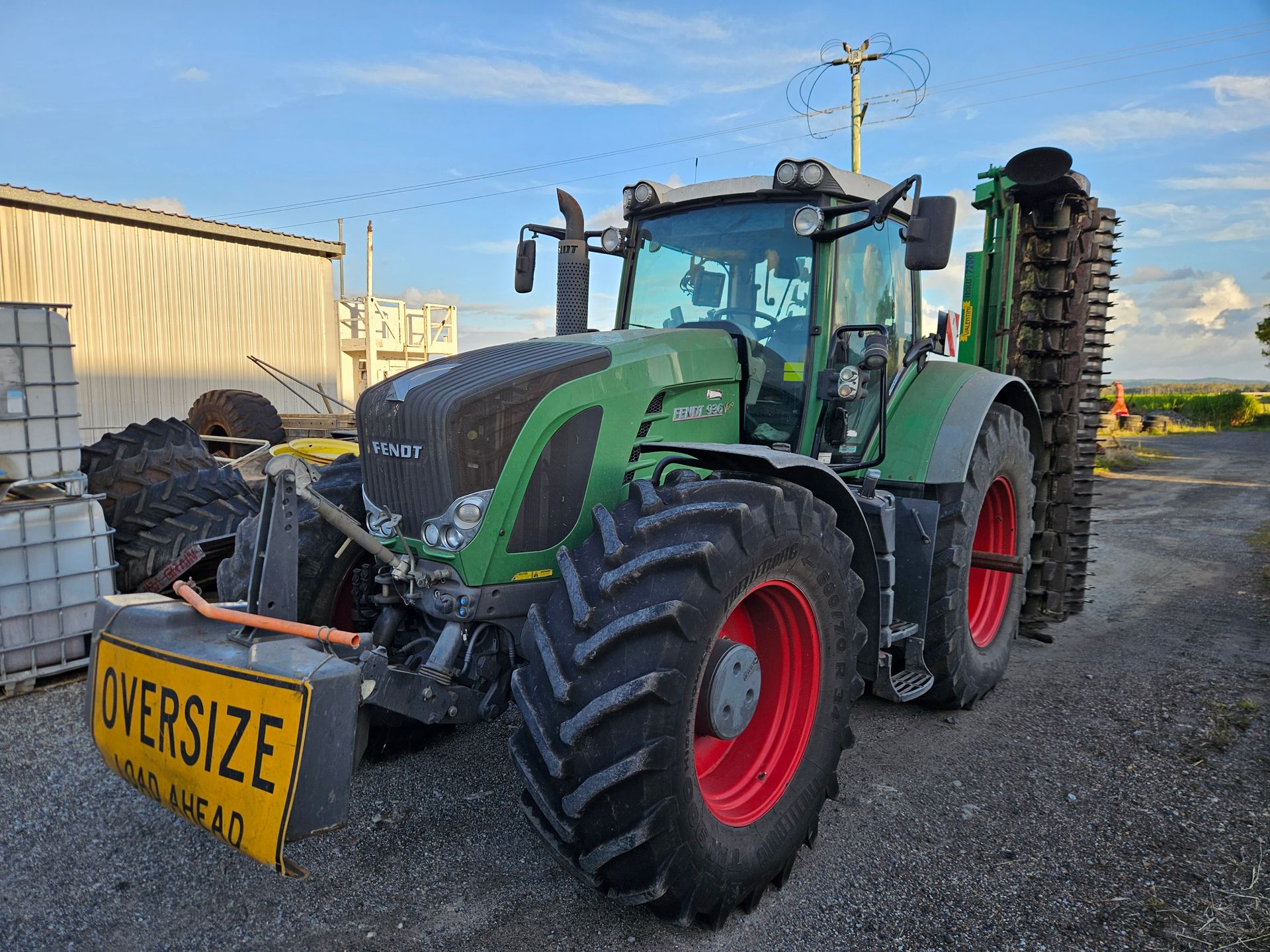 Green tractor with front-mounted oversized load sign parked on gravel near a farm building