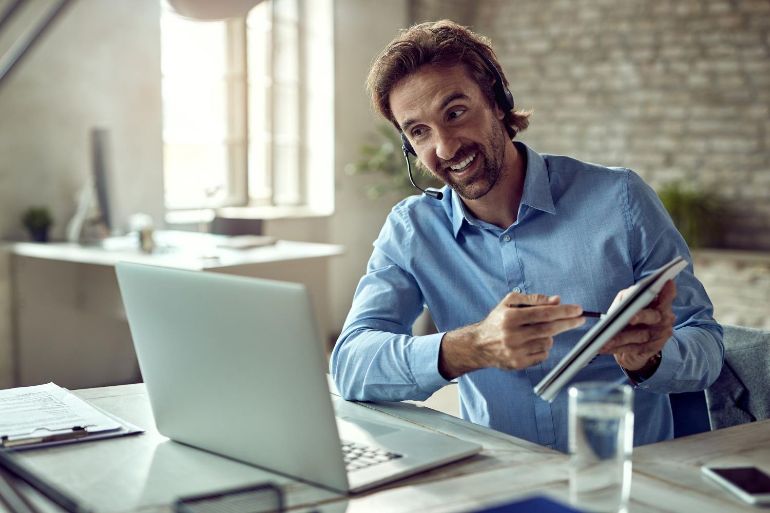 Man smiling at a desk, holding a tablet beside a laptop in a bright office