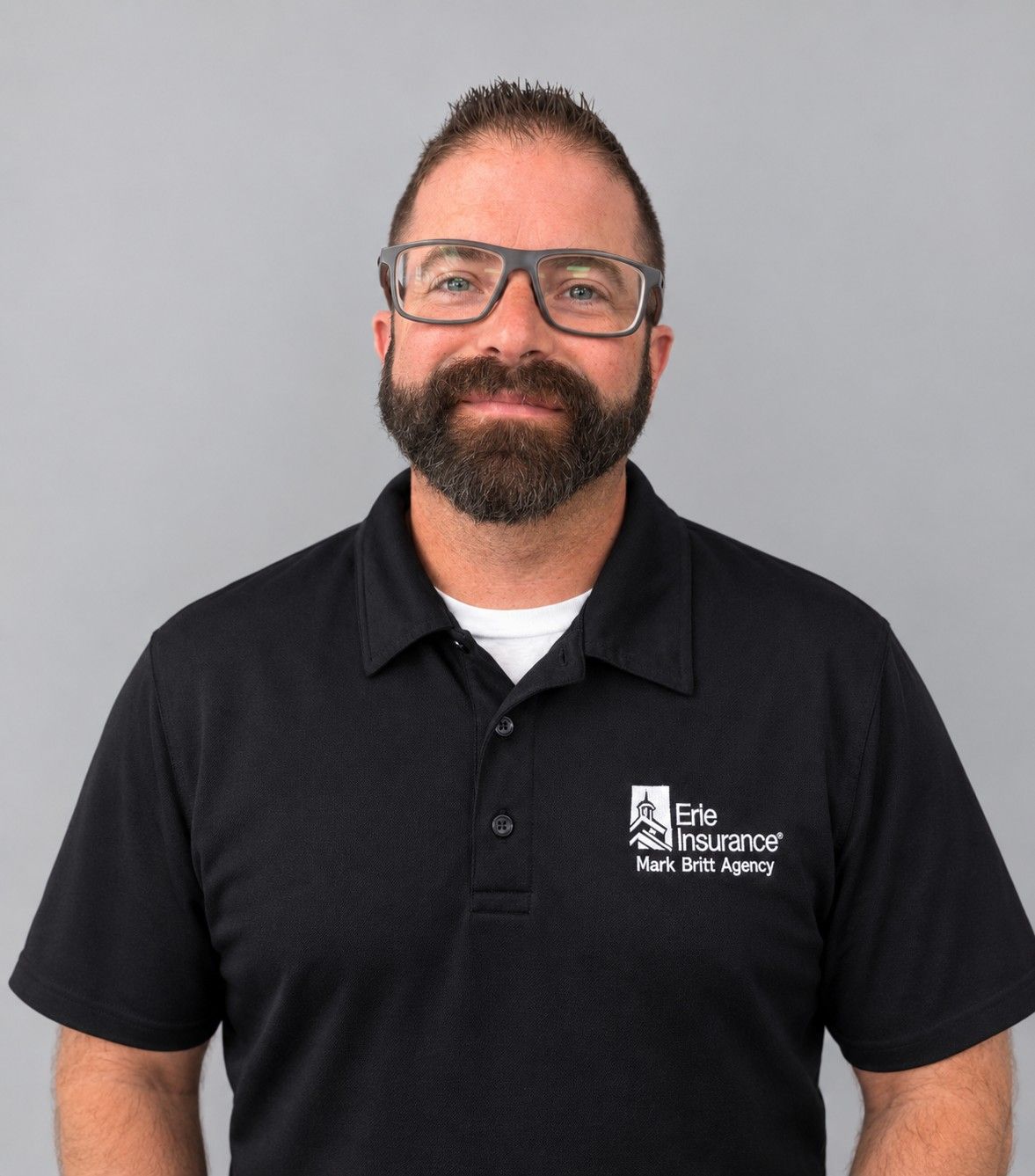 Man in black polo shirt and glasses, standing against a gray background, smiling slightly.