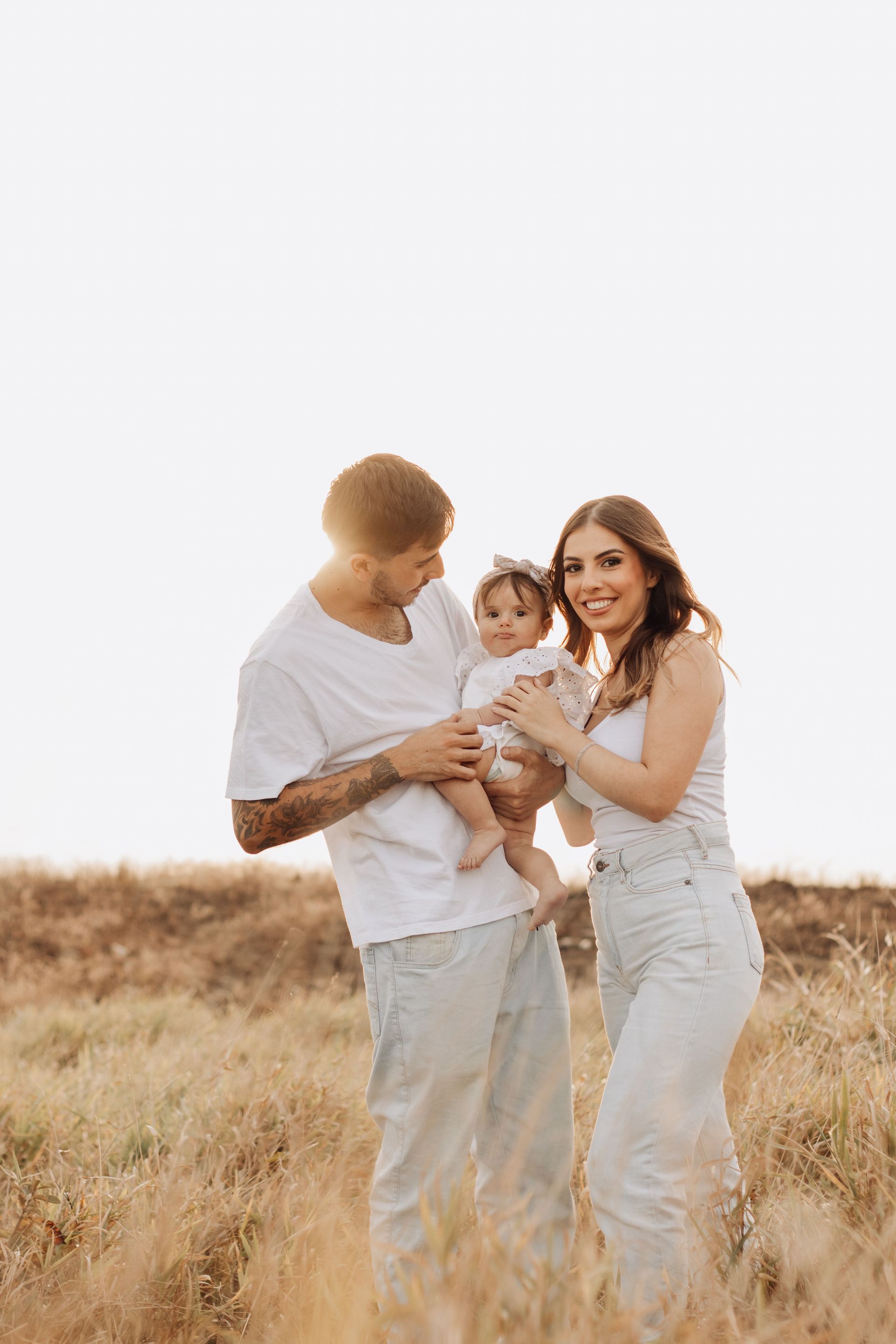 Family of three standing in a field, smiling and holding a baby in warm sunlight