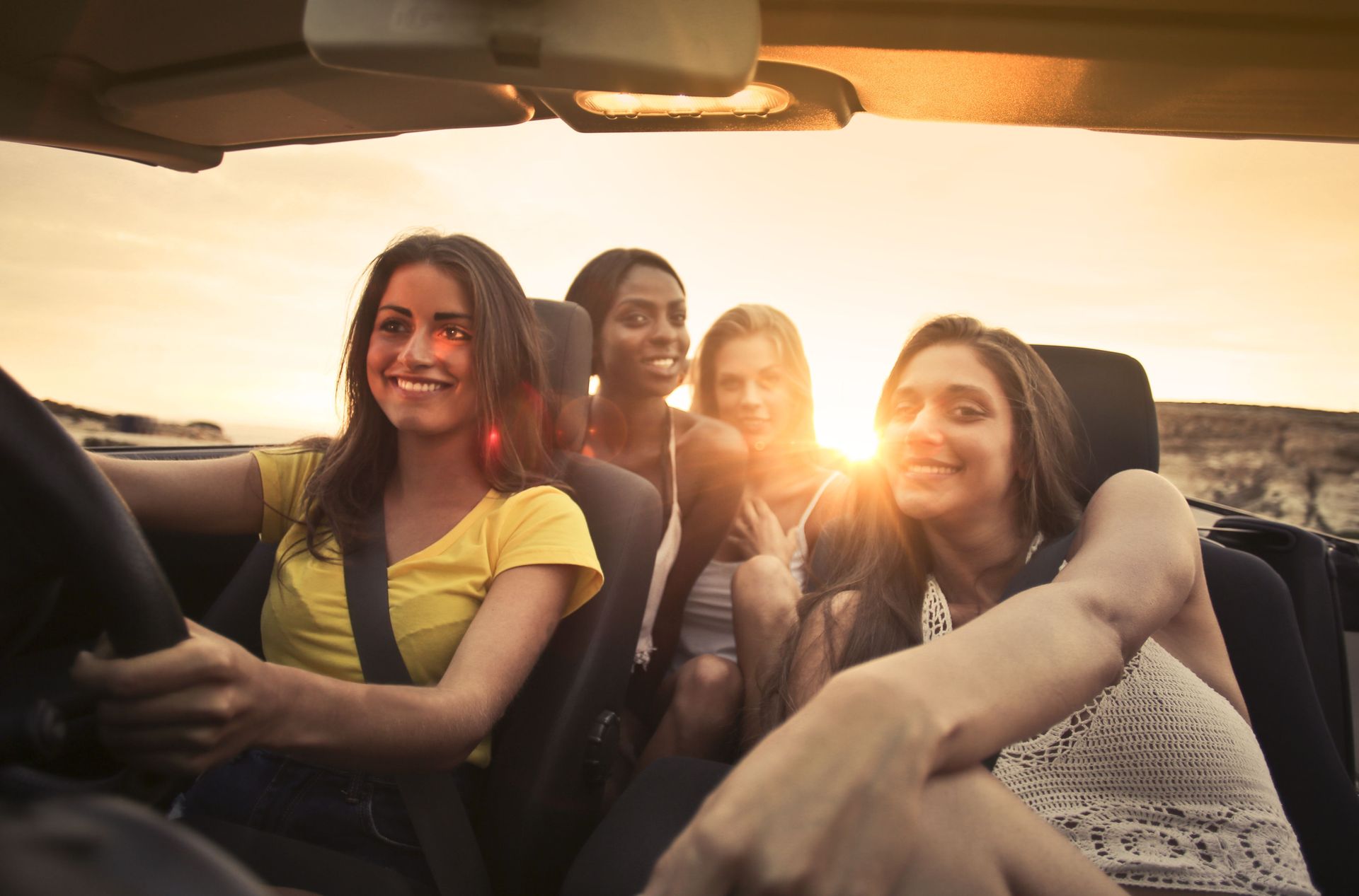 Four friends smile in a convertible at sunset, with warm sunlight and open road scenery.