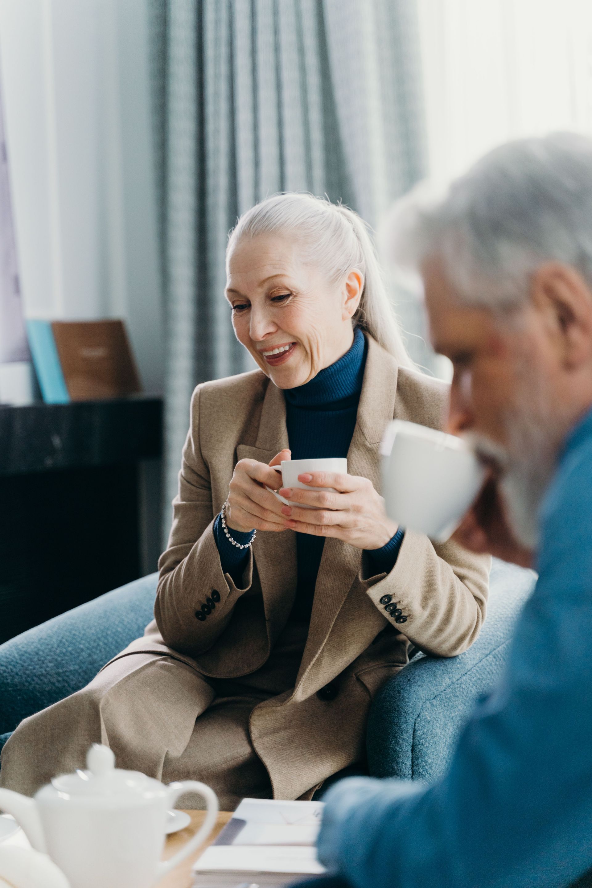 Two people in conversation, smiling and holding teacups in a bright living room.
