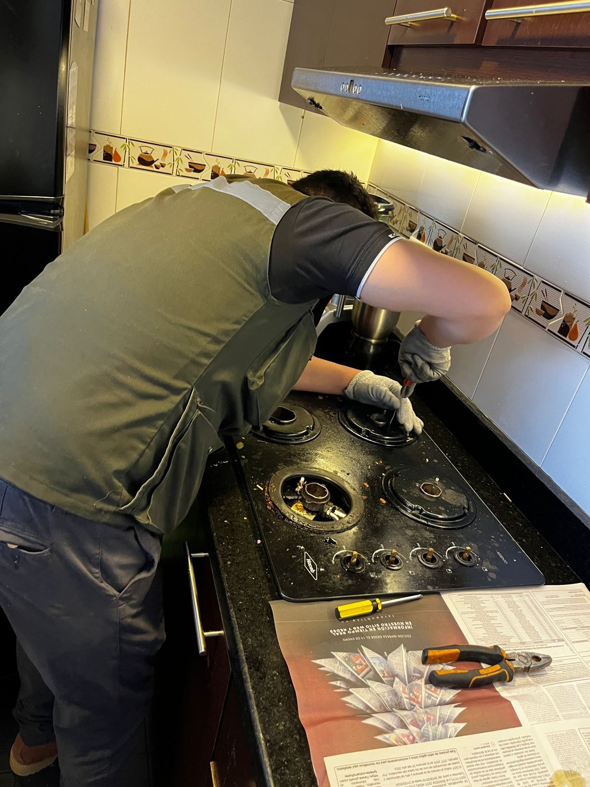 Una persona que viste un chaleco verde y guantes grises repara una estufa de gas negra en una cocina.