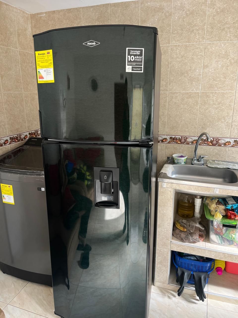 En una cocina hay un frigorífico negro con dispensador de agua, junto a una lavadora gris y un fregadero de hormigón.