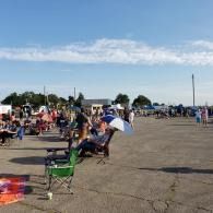 People sitting in folding chairs at an outdoor gathering on a sunny paved lot, with tents and a blue sky.