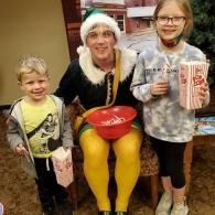 Person in elf costume attire seated between two children, holding a red bowl, with a popcorn bag nearby.