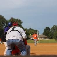 Baseball pitcher in orange on a dirt field, viewed from behind the catcher at home plate.