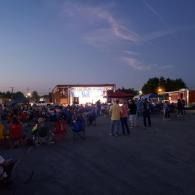 Crowd gathered at an outdoor evening event with a lit stage and food stalls under a twilight sky