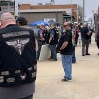 People in black biker vests gathering outdoors near a building, one man standing in jeans in the foreground
