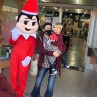 Woman holding a small child beside a person in an elf on the shelf costume at a store entrance