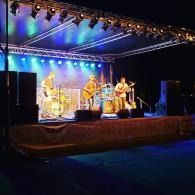 Outdoor stage with three musicians performing under blue and purple lights at night