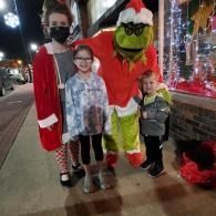 Three children posing with the Grinch in a Santa outfit on a city sidewalk at night