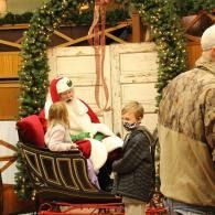 Santa seated in a sleigh speaks with two children at a holiday event decorated with greenery.