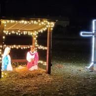 Outdoor nativity scene under string lights with figures in a straw manger and a glowing cross nearby