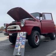 Red classic pickup truck with hood open and driver door ajar, parked on pavement with a display board leaning against it.