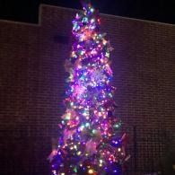 Tall Christmas tree covered in colorful lights glowing against a dark brick wall