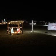 Illuminated nativity scene and glowing cross in a dark outdoor yard with a small sign nearby