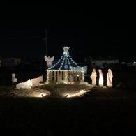 Nighttime outdoor display with a lit small structure, blue string lights, and nativity figurines nearby