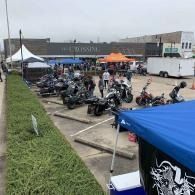 Outdoor motorcycle gathering in a parking lot, with many parked bikes, tents, and a blue canopy in the foreground