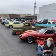 Classic cars lined up at an outdoor show, including a red roadster, with a seated man in the foreground.