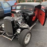 Black vintage hot rod with exposed engine and red interior, parked with driver door open.