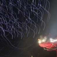Abstract firework light trails against a dark night sky, with white and red streaks near a bright glow.