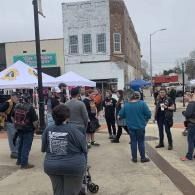 People gathered on a city sidewalk near white tents and a brick building, with some holding signs.