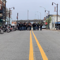 People standing across a city street, blocking the road near parked motorcycles and streetlights.