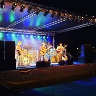 Band performing on a lit outdoor stage at night with blue and purple lights