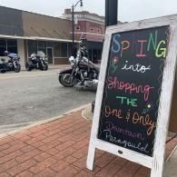 Sidewalk chalkboard sign outside storefront reads “Spring into shopping,” with motorcycles parked nearby.