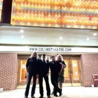 Four people standing outside a brick theater entrance under a bright marquee at night