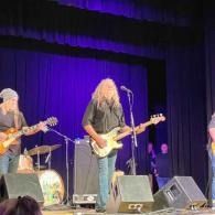 Live band performing on stage under purple lighting, with guitarists and drums visible.