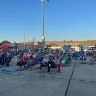 Crowd seated in folding chairs at an outdoor evening event in a parking lot under a blue sky