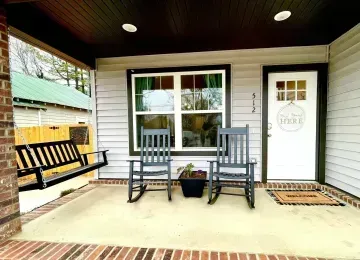 Front porch with two gray rocking chairs, black bench swing, white door, and potted flowers under a covered ceiling