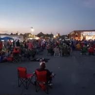Crowd gathered outdoors at dusk near a lit stage, with people seated in folding chairs on pavement.