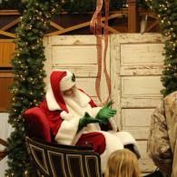 Santa in a red chair, wearing a red-and-white suit, sits in a festive holiday setting with garland and wood panels.