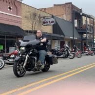 Motorcyclist riding past a row of parked motorcycles on a city street.