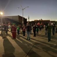 People standing spaced out in a parking lot at dusk, facing forward under streetlights.