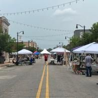 Outdoor market with white tents lining a closed street under string lights, people browsing booths