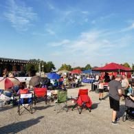 Outdoor crowd seated in folding chairs at a sunny event with tents and a red canopy.