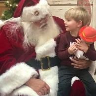 Santa holding a child with a red hat toy, both seated and smiling