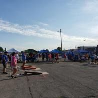 Outdoor cornhole games at a community fair, with blue tents and people gathered on a sunny day.