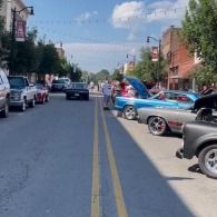 Classic cars lined up on a sunny street during a parade, with spectators and American flags.