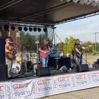 Four-piece band performing on an outdoor stage under a canopy, with “GET DOWN TOWN” festival banner along the front.
