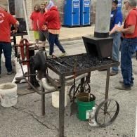 People in red shirts around a metal food-smoker cart outdoors with buckets and blue portable toilets nearby