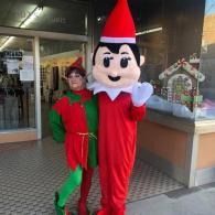 Elf on a shelf costumed person and a person dressed as an elf standing outside a storefront, waving.