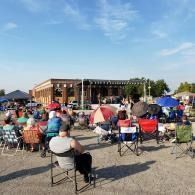 Outdoor concert crowd seated in folding chairs facing a brick stage under a clear blue sky